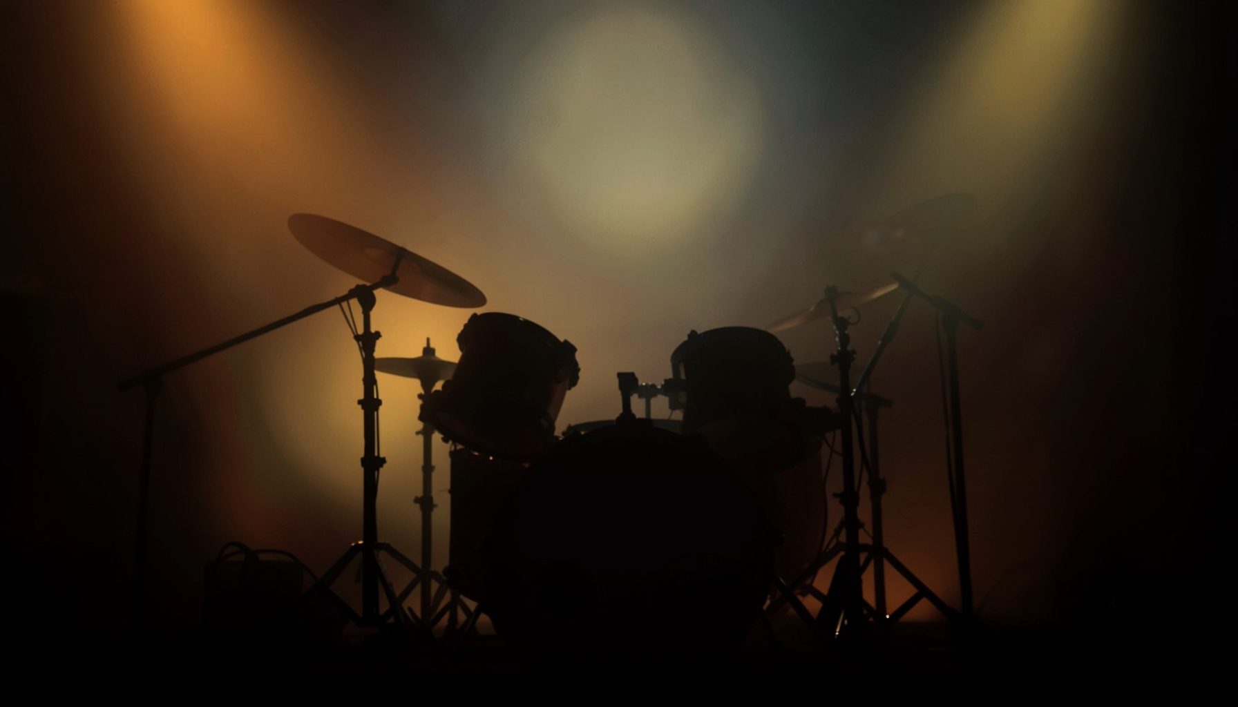 Warm stage-lit scene with drum kit silhouettes and deep amber and charcoal tones