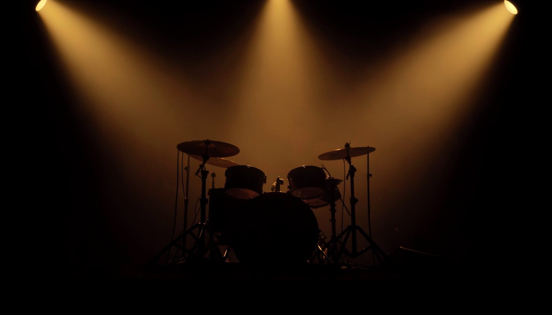 A dimly lit stage with drum kit silhouettes and warm amber spotlights, conveying the atmosphere of a live performance