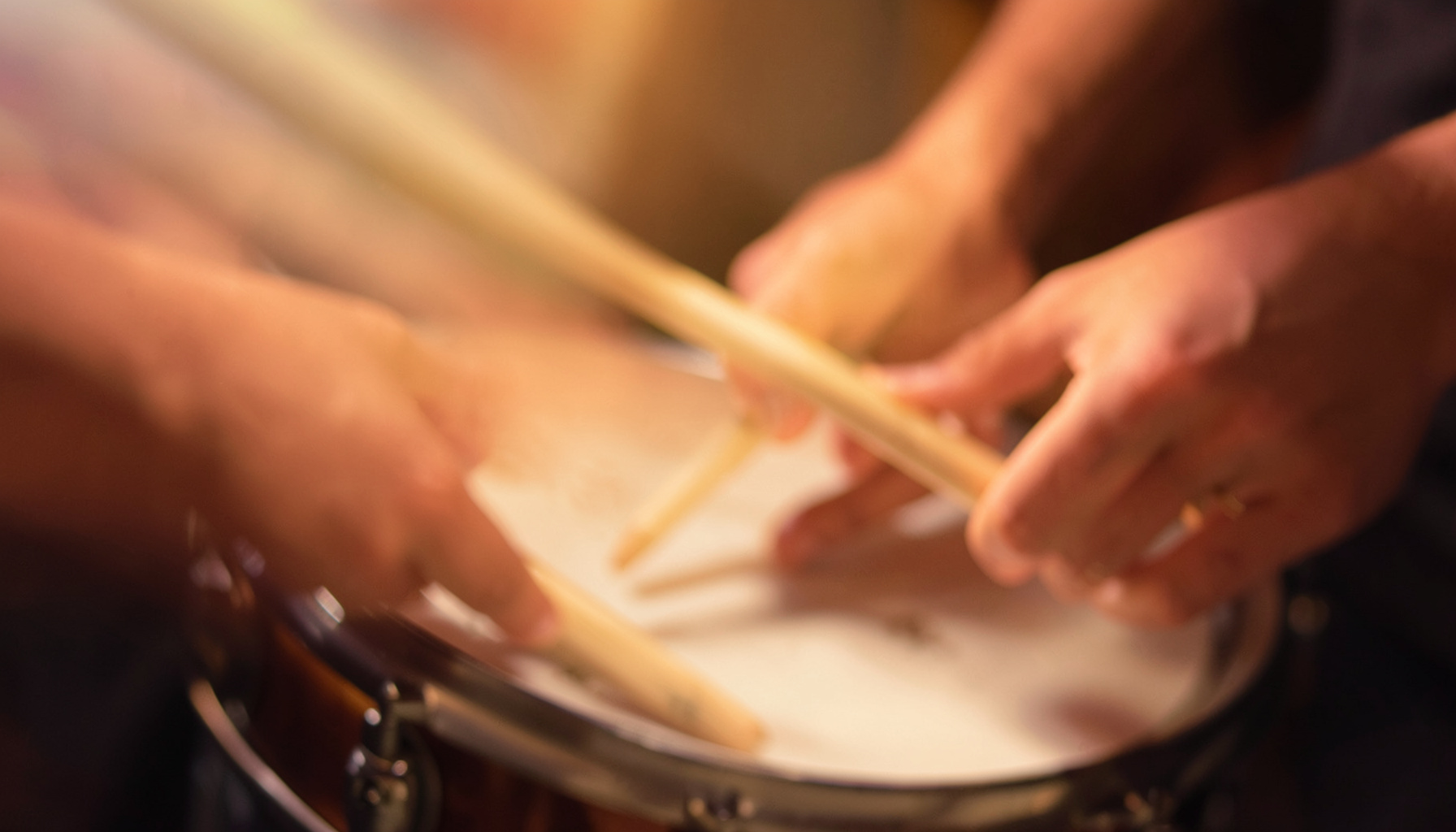 A close-up view of hands and drumsticks over a snare drum under warm studio lighting
